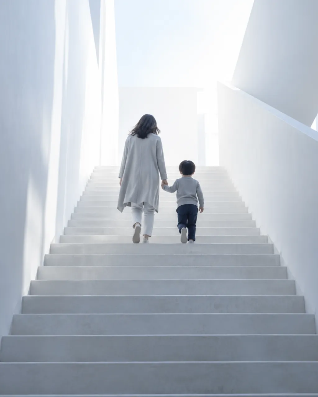 大人と子どもが手をつないで明るい階段を上る後ろ姿